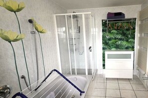Ein Badezimmer mit einer Glaskabinentrennung zur Dusche, einer Badewanne mit blauem Handtuchhalter und einem Waschtisch mit tropischem Blattentapete.