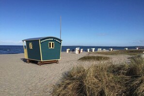 Ein Gebäude direkt am Strand mit einem Schild, auf dem 'STRANDKÖRBE & BOOTS' steht, vor einem sandigen Strand und einem wolkenlosen Himmel.
