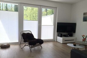 Ein Wohnzimmer mit Fernseher, einem Sessel, einem kleinen Tisch und einem Fenster mit Jalousien.