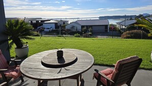 A patio with a round wooden table and chairs, a potted plant, and a view of houses and water in the background. - Omokoroa Sea View Apartment, stunning views, amazing sun rises, quiet spot (Ōmokoroa)