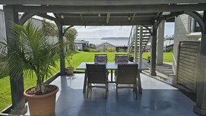 A covered patio with a table and chairs, a potted plant, and a view of a grassy area and buildings in the background. - Omokoroa Sea View Apartment, stunning views, amazing sun rises, quiet spot (Ōmokoroa)