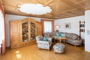 Ein Wohnzimmer mit Holzschrank, floralem Sofa, Pouf und einem kleinen Tisch.