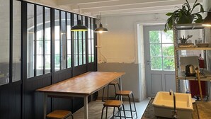 Una habitación con una larga mesa de madera, dos taburetes, un lavabo blanco y un estante con utensilios de cocina.