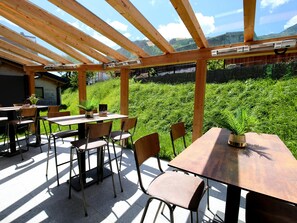 En uteservering med träbord och stolar, en pergola och utsikt över ett frodigt, grönt landskap.