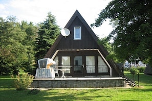 A triangular wooden house with a satellite dish, a small porch with a table and chairs, and a stone foundation. - Finnish hut Elisabeth (Gager)