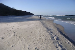 A sandy beach with footprints, a person walking, and a forested area in the background. - Holiday home in Rügen with 2 bedrooms (putgarten)