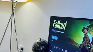 Un televisor mostrando la interfaz de Netflix con la serie 'Fallout' destacada, una lámpara, un globo terráqueo y un reposapiés blanco.