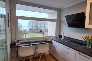 A modern kitchen with a large window, two white chairs, a black countertop, and a flat-screen TV mounted on the wall. - Ostseekieker-Kellenhusen (Kellenhusen)