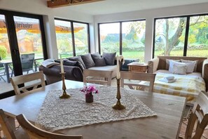 A room with a dining table, chairs, a sofa, and a bed. There are large windows with a view of greenery outside. - Romantic holiday home with sauna (Zossen)