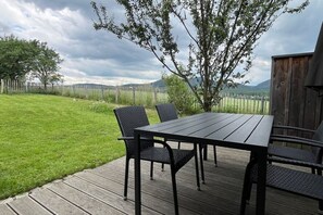 Eine Terrasse mit Tisch und Stühlen, umgeben von einem Holzzaun, mit Blick auf ein Wiesenfeld und ferne Hügel.