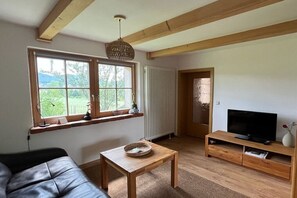 Ein Wohnzimmer mit Sofa, Holztisch, Fernseher auf Holzständer, Fenster mit Blick auf Grünflächen und Heizkörper.