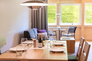 A dining table set with wine glasses, plates, and a bottle of wine, with a view of greenery outside. - Rössle Apartments Type 3 (Fontanella)