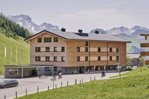 A multi-story building with a brick facade, a covered entrance, and a view of mountains in the background. - Rössle Apartments Type 3 (Fontanella)