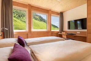 A room with two beds, a desk, a chair, a television, and large windows with a view of a green landscape. - Rössle Apartments Type 3 (Fontanella)