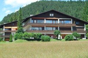 A multi-story building with balconies, surrounded by greenery and a forested hill.