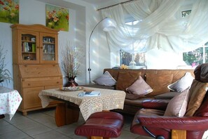 Ein gemütliches Wohnzimmer mit einer Holzkabine, einem Tisch mit einer Spitzentischdecke, einem Ledersofa und einem großen Fenster mit transparenten Vorhängen.
