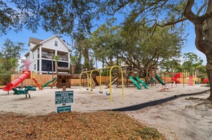 A playground with various play equipment including slides and swings, surrounded by trees and a building. - Admirals Row, Ocean Front Villa-Hilton Head Beach and Tennis (Hilton Head Island)