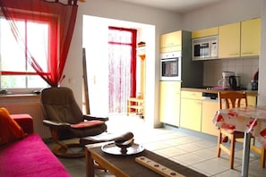 Ein Wohnzimmer mit einer Couch, einem Sessel, einem Esstisch und einer Küchenzeile mit gelben Schränken und einer Mikrowelle.