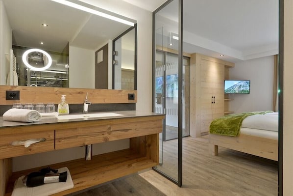 Ein modernes Hotelzimmer mit einem Holz-Waschtisch, einem großen Spiegel, einem Flachbildfernseher und einem Bett mit grüner Decke.