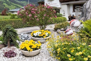 Ein Garten mit unterschiedlichen Pflanzen und Blumen, ein Gebäude mit einem Schild und eine gebirgige Landschaft im Hintergrund.