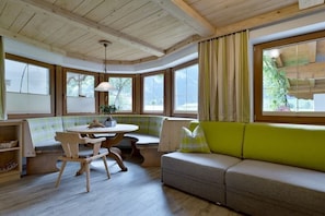 Ein gemütliches Zimmer mit Holzbalkendecke, einem runden Tisch, einer Couch mit Kissen und großen Fenstern.