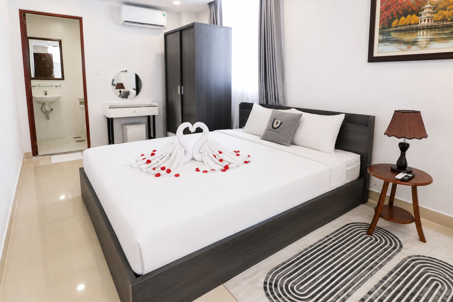 A hotel room with a large bed, a bedside table with a lamp, a wardrobe, a bathroom, and a painting on the wall.