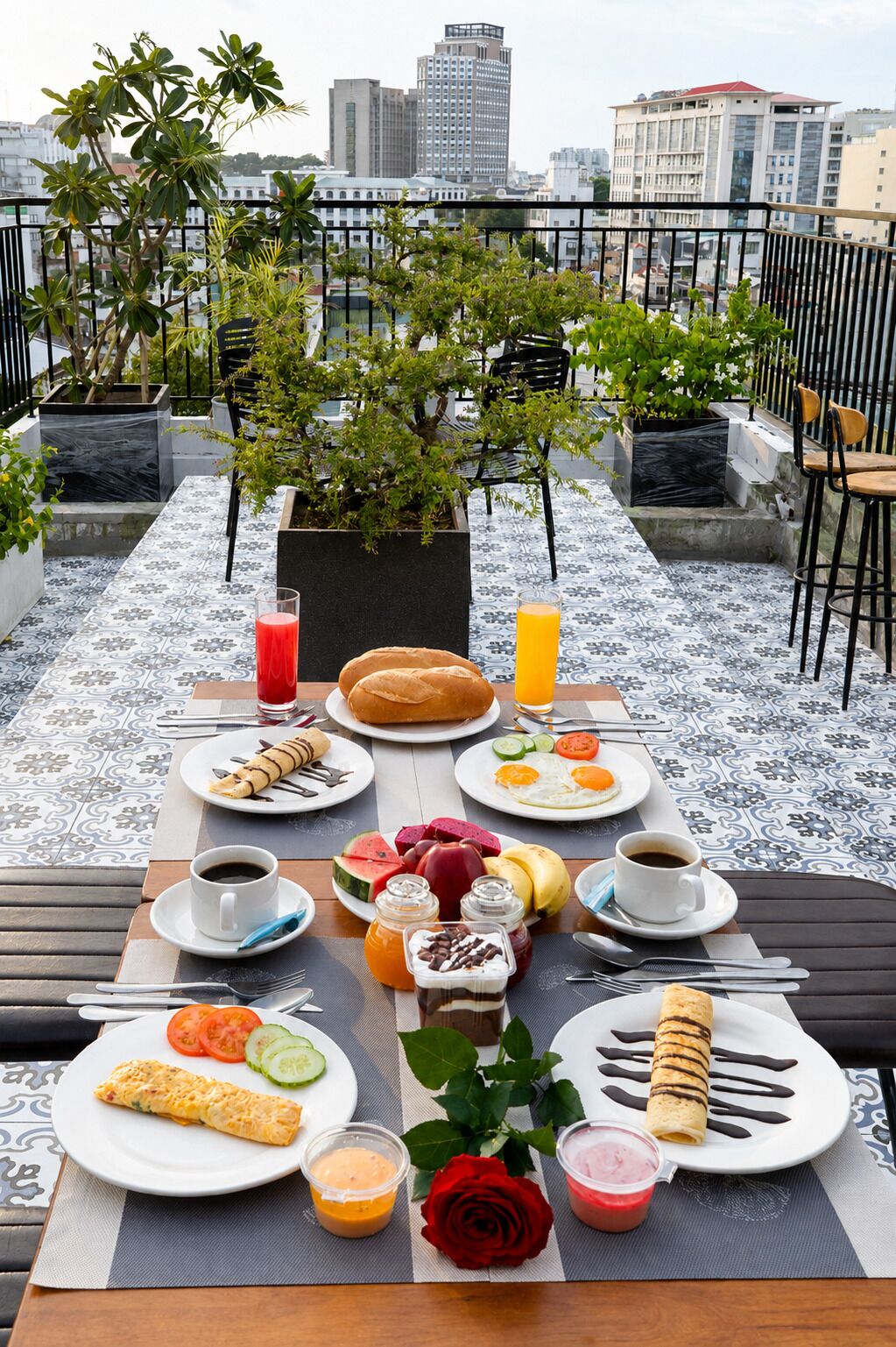 A rooftop dining setup with a city view, featuring a table with various breakfast items, plants, and a rose.