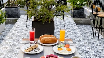 Un espacio para comer en la azotea con vistas a la ciudad, que incluye una mesa con diferentes opciones de desayuno, plantas y una rosa.