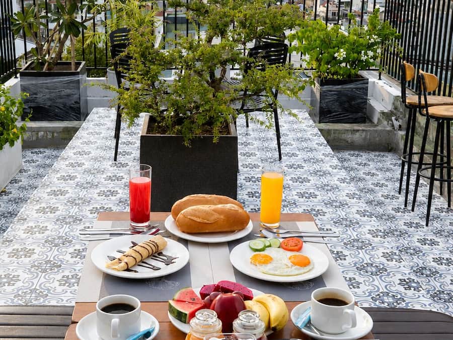 Un espacio para comer en la azotea con vistas a la ciudad, que incluye una mesa con diferentes opciones de desayuno, plantas y una rosa.