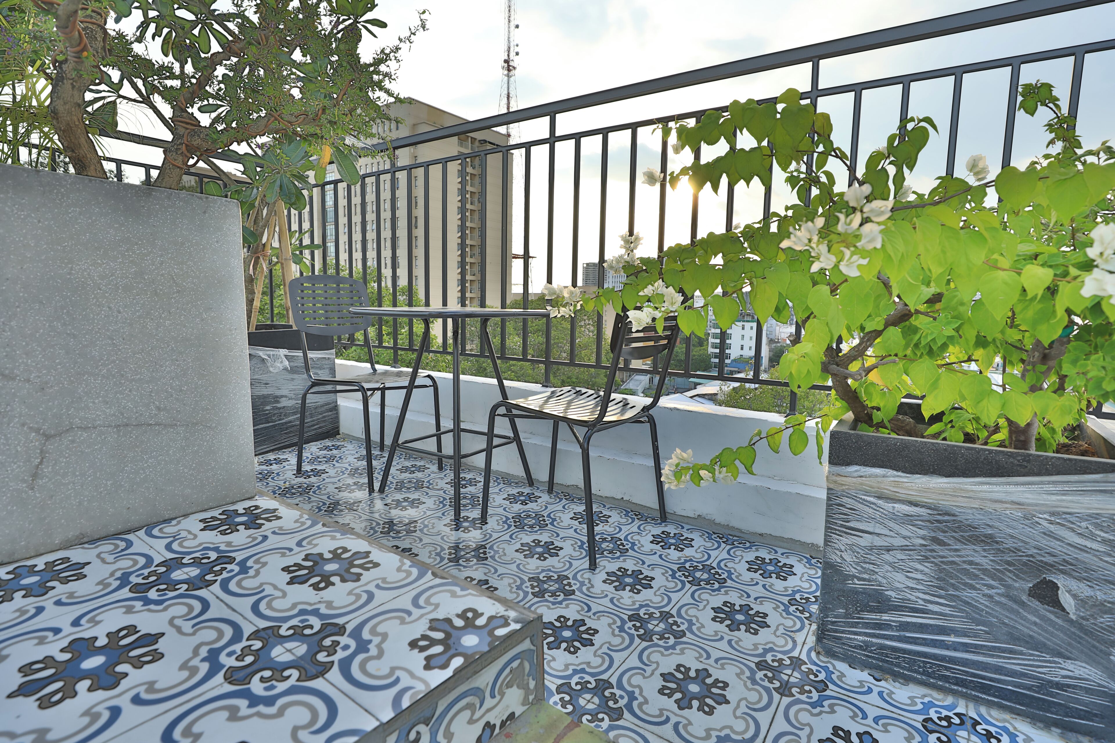 A balcony with patterned floor tiles, two metal chairs, a potted plant, and a view of a building in the background.