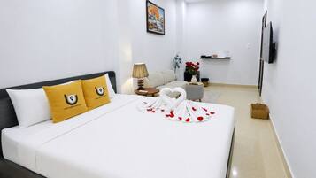 A hotel room with a bed, a nightstand, a lamp, a vase with red flowers, a wall-mounted TV, and a painting on the wall.