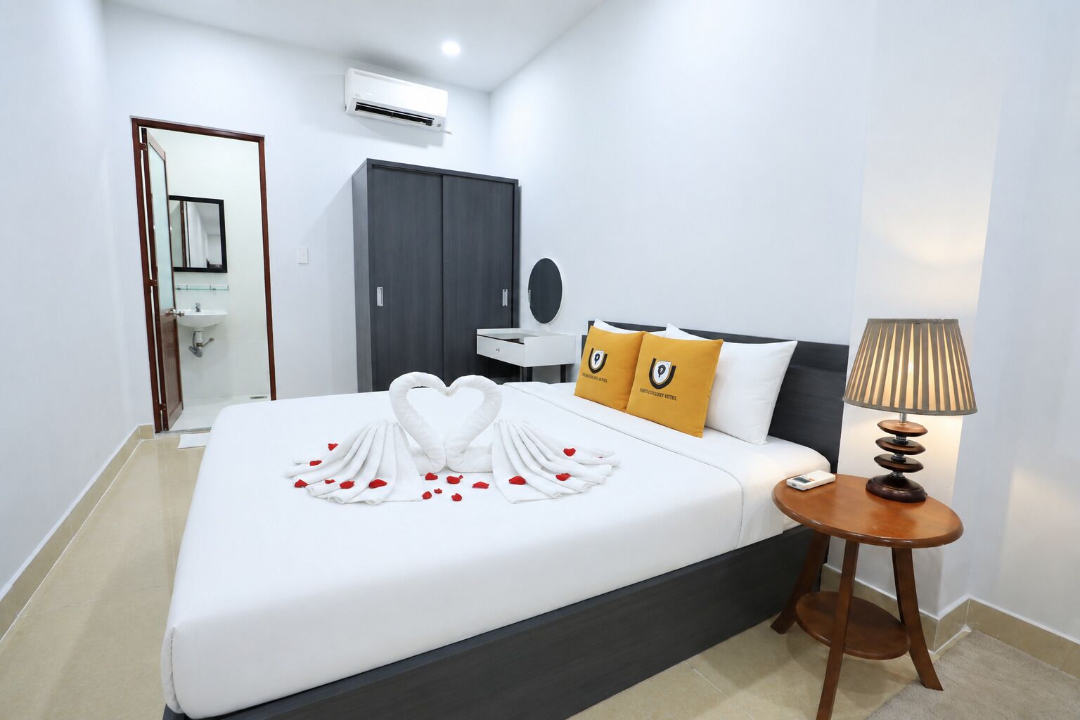 Habitación de hotel con cama, mesita de noche con lámpara, armario y baño visible a través de una puerta abierta.