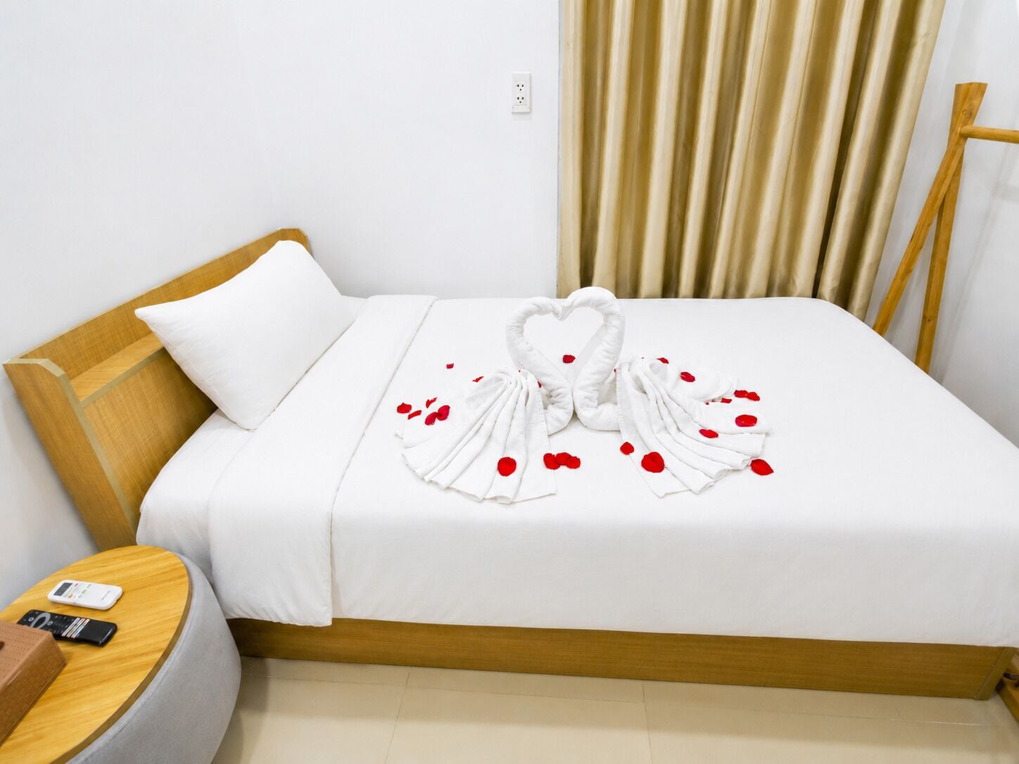 A hotel room with a neatly made bed, a wooden headboard, a bedside table with a remote control, and a power outlet on the wall.