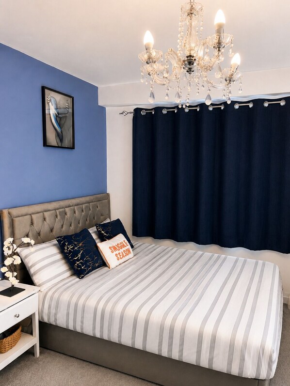 A bedroom with a tufted headboard, striped bedding, a chandelier, a framed picture, and dark curtains.