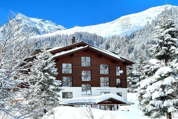A wooden building with multiple windows, surrounded by snow-covered trees and mountains. - 4 in the Bohemia House (Gargellen)