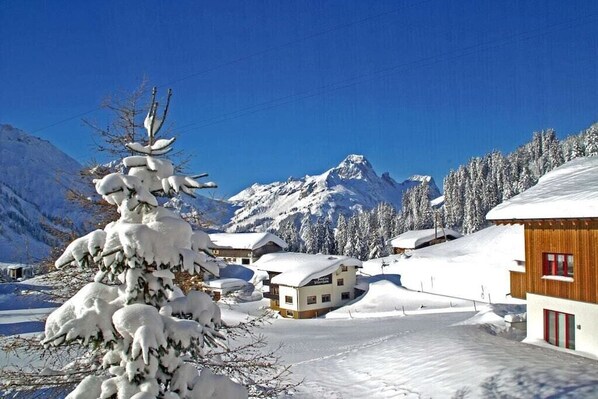 A snow-covered mountain village with chalet-style buildings, a clear blue sky, and a prominent mountain peak in the background. - Rothorn - App. Braunarl (Schröcken)