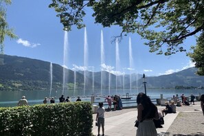 Blick auf ein Seeufer mit mehreren Springbrunnen, einem gepflasterten Weg und Menschen, die die Landschaft genießen.