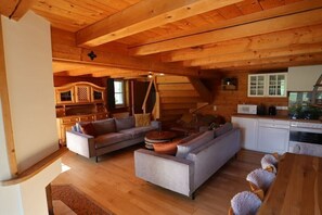 Ein Blockhaus-Interieur mit Holzbalkendecke, einer Sofagarnitur, einem Esstisch und einer Küchenzeile.