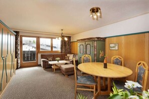 Ein Zimmer mit einer Sofagarnitur, einem Esstisch mit Stühlen, einem Schrank und einem Fenster mit Vorhängen.