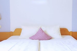Ein Bett mit weißer Bettwäsche und zwei Kissen, eines rosa und eines weiß, in einem Zimmer mit hellen Wänden.