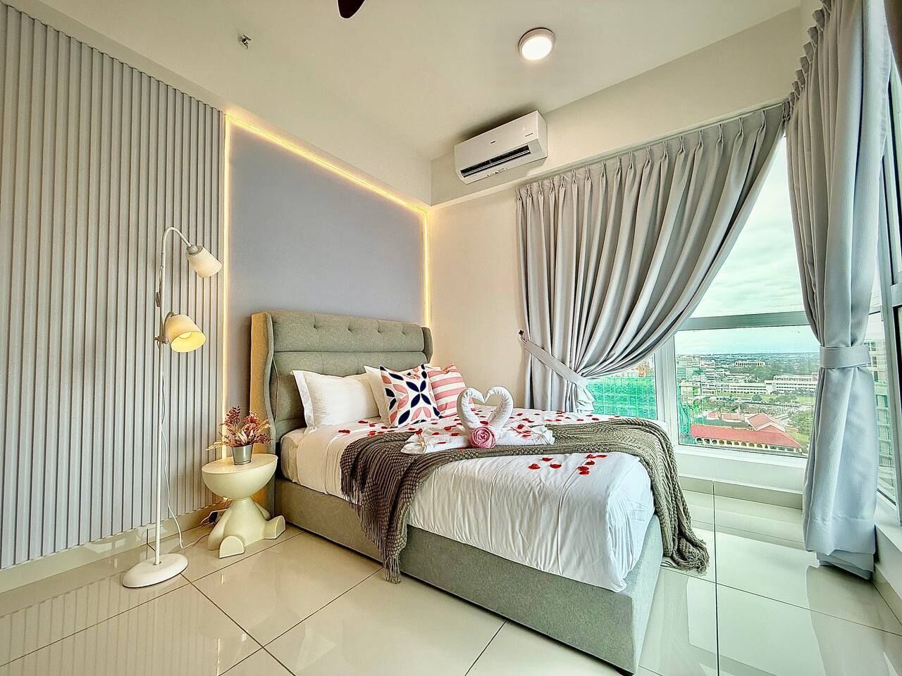 A modern bedroom with a large bed, a bedside table with a lamp, and a view of a cityscape through the window.