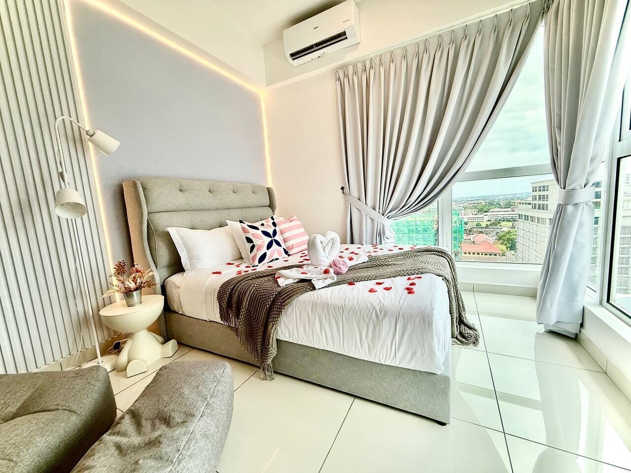 Un dormitorio moderno con una cama grande, una mesita de noche, una silla y vistas a la ciudad a través de amplias ventanas.