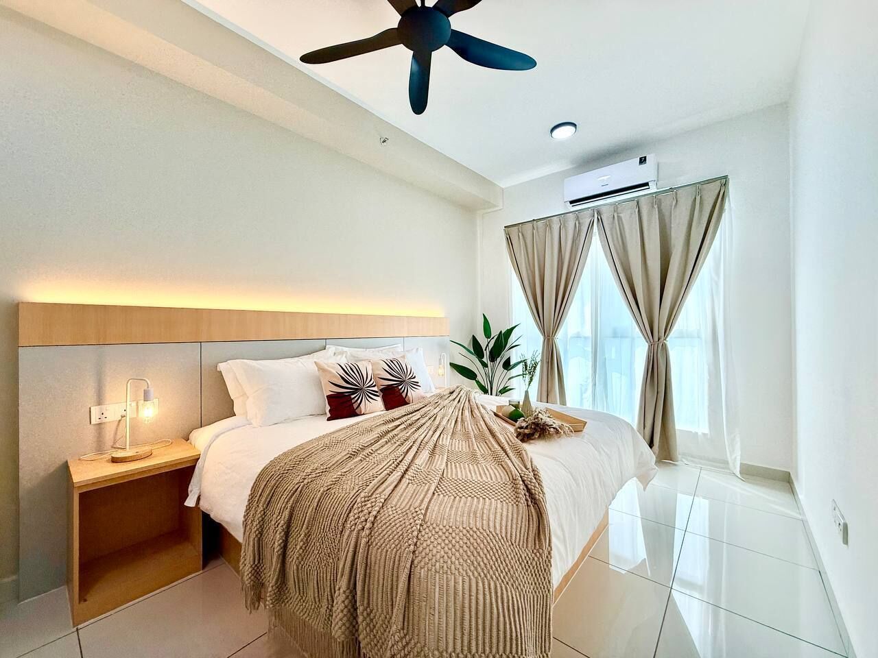 Habitación de hotel moderna con cama, mesita de noche, lámpara, ventilador de techo, aire acondicionado, cortinas y una planta en maceta.
