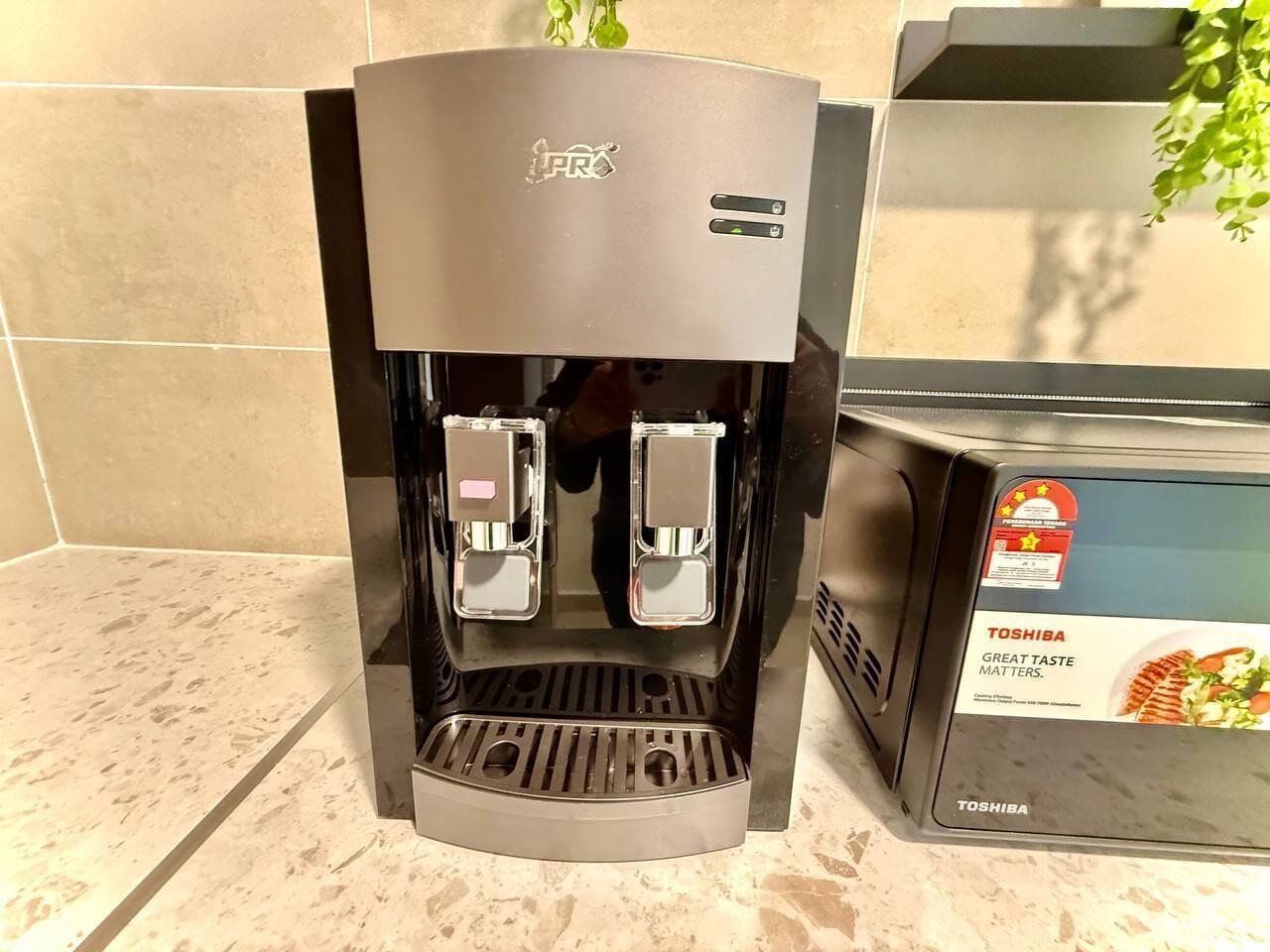 A coffee machine with two dispensers, a Toshiba branded box, and a tiled wall.