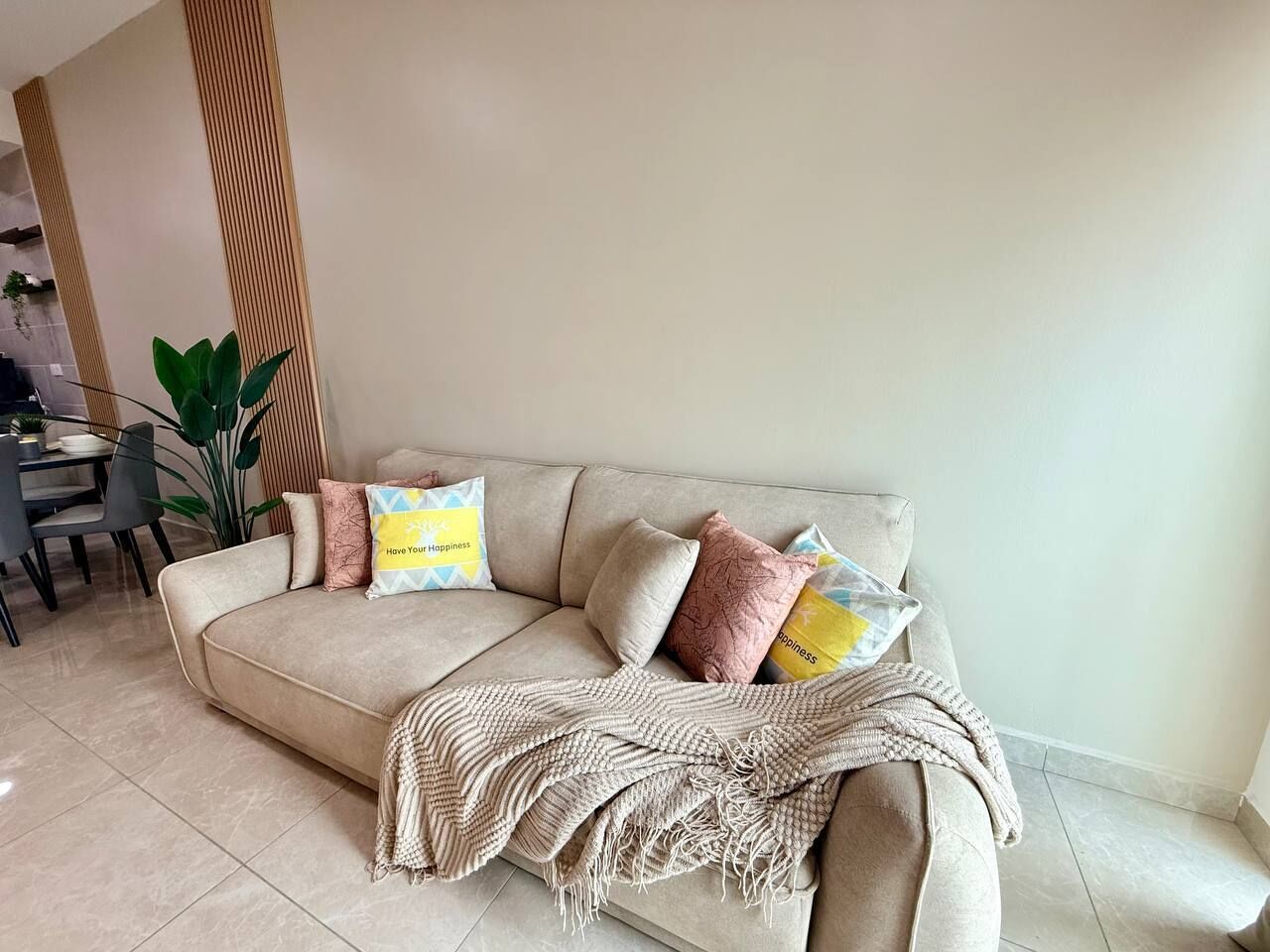 Een beige bank met meerdere kussens, een geweven plaid en een eethoek met stoelen en een tafel.