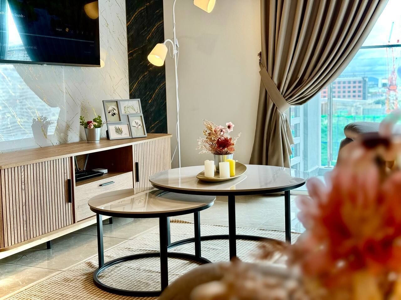 A modern living room with a round table, a TV mounted on the wall, and a city view through large windows.