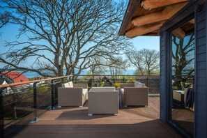 A deck with wicker furniture, a glass railing, and a view of trees and a body of water. - Pilot houses Rügen - Luxury Villa 4 (Breege)