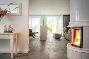 A modern living room with a fireplace, a dining area, and a view through a large window. - Pilot houses Rügen - Luxury Villa 4 (Breege)