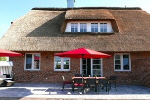 Ein Gebäude mit strohgedecktem Dach und einer Ziegelaußenwand, einem kleinen Fenster und einer Terrasse mit Tisch und Stühlen.