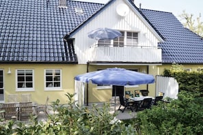 Ein Haus mit blau gefliestem Dach, einem Balkon mit weißer Geländer und einer Terrasse mit Tisch und Stühlen unter einem blauen Sonnenschirm.